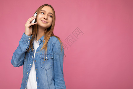 照片中 迷人 积极的年轻金发女性身穿休闲蓝色牛仔衬衫 在粉红色背景中显得格格不入 手牵着手 用手机看着旁边背景
