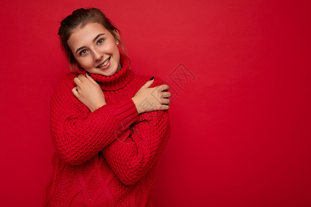偎穿着温暖的红色毛衣的迷人可爱微笑的年轻女性被隔离在红色背景墙上 手放在胸前看着相机头发拥抱空间女士背景