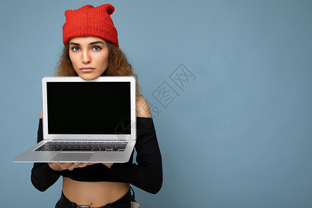 浅蓝色上衣特写肖像的滑稽美丽的深色金发女人拿着笔记本电脑看着相机穿着黑色裁剪上衣和红色和橙色的 dorag 隔离在浅蓝色的墙壁背景网络监视背景