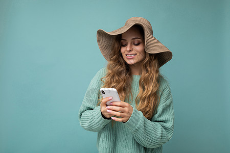 身着便衣的美丽年轻女子透过手机看移动屏幕 在互联网上背景冲浪时与外界隔绝地站立卷曲神器互联网女性消息帽子网络机动性电话蓝色背景图片