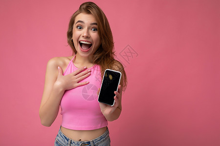 三品一标照片中 身穿粉红色上衣的美丽 快乐的金发年轻女子在粉红色背景中与世隔绝 手握空白空间 并展示带有空显示屏的手机 用于模拟看相机短背景