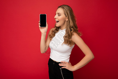 三品一标惊讶的美丽年轻金发女性穿着白色 T 恤 与红色背景隔离 复制空间拿着智能手机 手拿手机 屏幕空白 用于模拟查看设备显示屏背景
