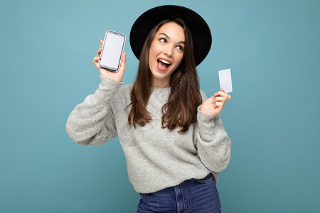 蓝色背景中 身穿黑色帽子和灰色毛衣的迷人 快乐的黑发年轻女性手持信用卡和手机的照片 显示空白 供模特看向侧面银行货币互联网电子展背景图片