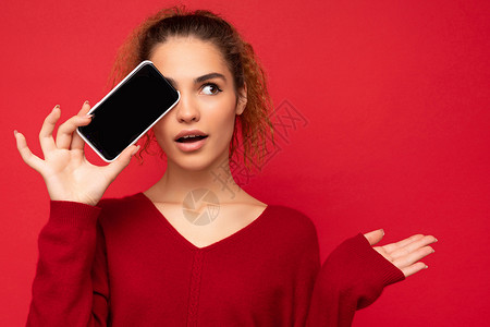 三品一标身着深红色毛衣的快乐有趣的年轻女性 被隔绝在红背景上 持有智能手机并展示移动电话屏幕及影印空间 供人们看侧面时使用背景
