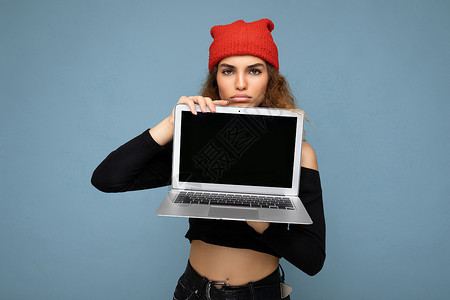 浅蓝色上衣特写肖像的滑稽美丽的深色金发女人拿着笔记本电脑看着相机穿着黑色裁剪上衣和红色和橙色的 dorag 隔离在浅蓝色的墙壁背景社交网页背景