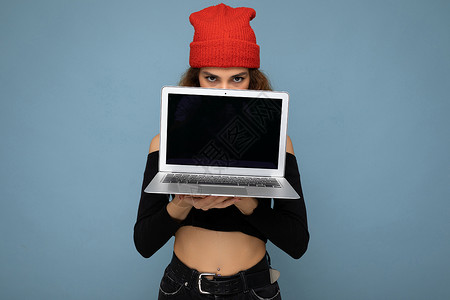 浅蓝色上衣特写肖像的滑稽美丽的深色金发女人拿着笔记本电脑看着相机穿着黑色裁剪上衣和红色和橙色的 dorag 隔离在浅蓝色的墙壁背景互联网网背景