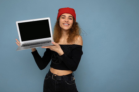 浅蓝色上衣特写 滑稽微笑 快乐 美丽的深色金发年轻女子手持笔记本电脑 看着相机 身穿黑色裁剪上衣 红色和橙色 do-rag 与浅蓝色墙壁背背景