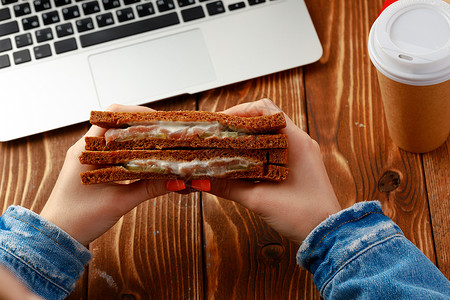 一名用笔记本电脑在工作台上方手持三明治的妇女的手键盘职场小吃咖啡女性午餐食物杯子办公室桌子背景图片