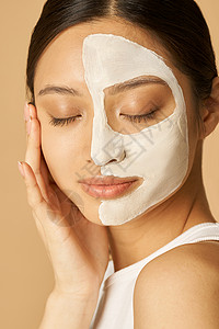 涂润肤霜放松的年轻女性 在半张脸上涂上面膜 接受水疗护理 在米色背景下闭着眼睛对着镜头摆姿势背景