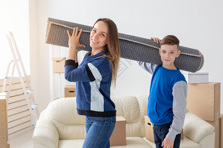 微笑的白人母亲和迷人的儿子在新客厅里肩上扛着折叠地毯 打算把它铺在新公寓里背景图片