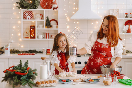 准备提供帮助新年厨房的妈妈和女儿 一起为圣诞饼干准备点钱吃吧围裙帮手家庭面团帮助女士拥抱桌子小姑娘房子背景