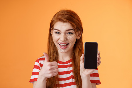 充满自信的红头发女人推荐智能手机应用程序 显示电话展示 拇指像批准标志 笑得面带微笑 建议良好的网上购买 移动广告等内容促销橙子背景图片