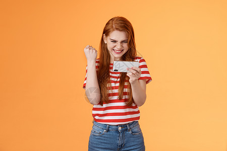 高兴的幸运红发女玩家红发 拳头满意 说是兴奋的凝视显示 水平握住智能手机 通过硬关卡玩很棒的游戏使用应用程序 取得胜利女士技术学背景图片