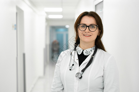 女医生画像站在医院走廊里的女医生 一位戴眼镜的年轻女医生和一件白大衣在现代诊所摆姿势的画像 看照相机的骄傲的职业女性医生治疗师风镜员工外套背景