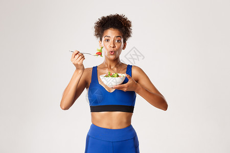 健康的生活方式和体育理念 适合的非洲-美体健美女性在吃沙拉时感到惊讶 站在白种背景之上背景图片