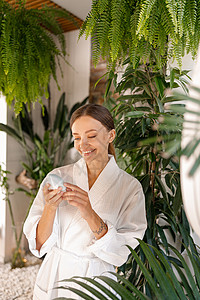 白色浴袍浴袍中快乐的年轻女子拿着硅胶刷刷清洗装置 同时在浴室里用植物装饰美容习例背景