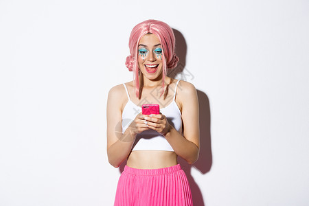 穿着粉红色假发参加派对 万圣节庆祝活动 站在白背景上站立的喜讯中 在手机上看着快乐新闻的惊讶女孩的肖像背景图片