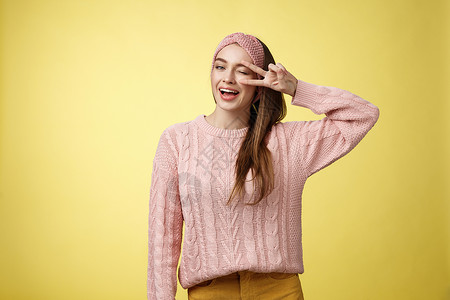 专享福利身穿粉色针织毛衣 头戴头带 眨眼轻浮可爱 在眼睛上展示胜利或和平标志 在黄墙上感到兴奋和快乐的快乐快乐魅力的年轻欧洲女性女朋友享背景