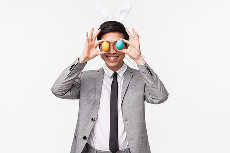 穿西装的兔子欢乐节 人与庆祝概念 喜悦式画像 由穿着灰色西装和领带 把彩蛋蒙在眼睛上并微笑 享受复活节派对的风趣 嬉戏和充满热情的亚洲男子组背景