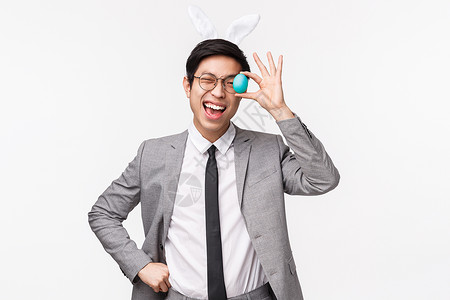 穿西装的兔子欢乐节 人与庆祝概念 喜悦的快乐有趣的年轻亚洲男子穿着灰色西装和领带 眨眼并展示彩蛋以享受复活节 戴兔子耳朵背景