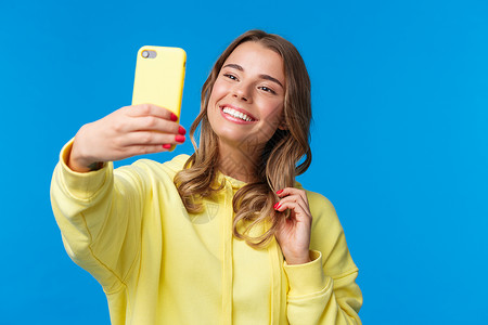 特写肖像温柔可爱的金发女孩穿着黄色连帽衫 手持手机 用智能手机自拍 在应用程序中添加照片滤镜 站立蓝色背景背景