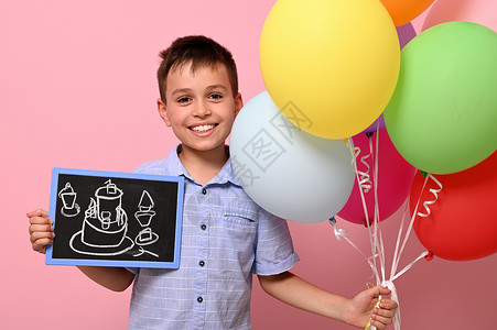 撑起黑板的气球可爱的男孩手持多色彩色彩色气球 另一手拿着一张纸板和一份生日蛋糕 粉红色背景与复制空间隔绝背景