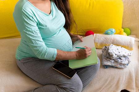 保活机制可爱的孕妇用笔记本为妇产医院写装箱单准备袋子 怀孕期间的年轻 Ledy 在家里的沙发上玩得很开心 母性 医学健康和护理理念婴儿保背景