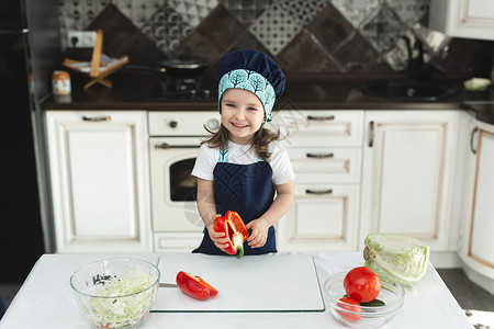 一个穿着围裙的孩子 厨房里一个厨师的帽子 在准备沙拉 吃红胡椒和大笑图片
