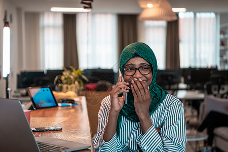 微笑的年轻女子谈论智能手机工作在笔记本电脑上看远距离思维 快乐的非洲裔美国客户在家庭办公室使用现代小工具手机和电脑一代视频媒体商背景图片