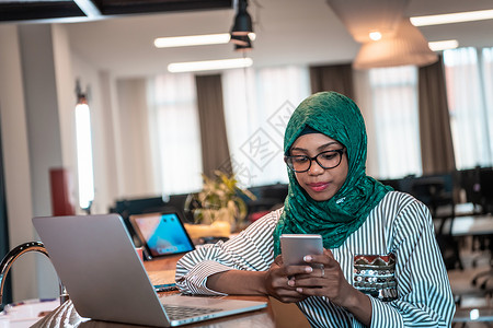 微笑的年轻女子谈论智能手机工作在笔记本电脑上看远距离思维 快乐的非洲裔美国客户在家庭办公室使用现代小工具手机和电脑趋势咖啡厅媒体背景图片