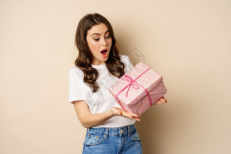 庆祝活动和假期的概念 快乐的年轻女人拿着用粉色盒子包裹的礼物 收到礼物 站在米色背景上 看起来既惊讶又惊讶乐趣女孩喜悦标识促销微背景图片