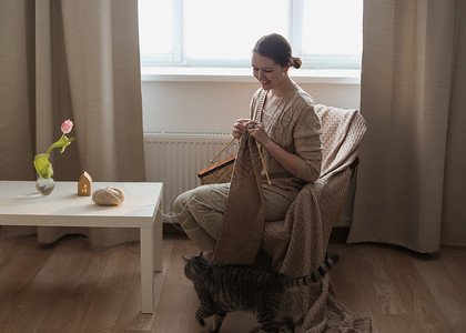 家里客厅的猫女人从羊毛纱和可爱有趣的小猫编织 在舒适的阳光明媚的房间在家里客厅格子朋友窗户家庭微笑闲暇公寓针织床单背景