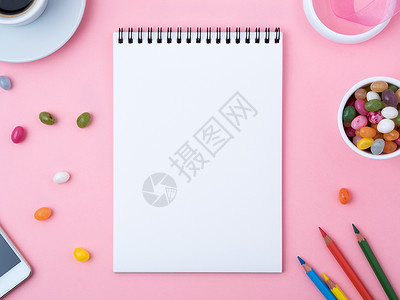 白纸糖果边框打开笔记本 上面有一张干净的白纸 焦糖 棒棒糖 手机 蜡笔 一杯咖啡 粉红色亮桌上的装饰品 女孩的创造力 计划和梦想的工作场所背景