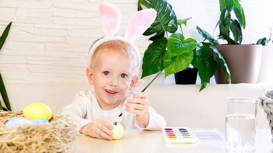 画彩蛋的孩子一个小兔子耳朵里的小男孩 热情地用油漆给复活节鸡蛋涂彩画 宗教假日概念 你自己做 家庭休闲 东边猎鸡假期幼儿园戏服教育篮子画笔孩背景