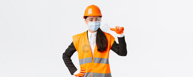 装修宣传折页Covid19 在企业 建设和预防病毒概念方面的安全协议 自信的亚洲女工程师戴着头盔和面罩指着自己 宣传个人协助女孩商业快乐建筑背景