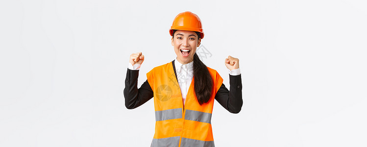 成功获胜的亚洲女工程师举起拳头 欢呼雀跃 戴上安全帽和夹克 战胜胜利 及时完成建设 站在满意的白色背景下房地产女孩人士成功快乐领背景图片