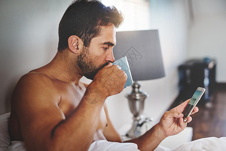 一边喝咖啡一边查看他的消息 一个英俊的年轻人在家里的床上喝咖啡时检查他的消息的裁剪镜头背景图片