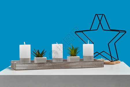 描边星星白色桌子上放着盆栽绿色植物 三根大蜡烛和一根小烛台 木架上的铁星形式 蓝色背景 特写背景