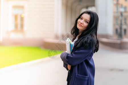 美丽的微笑女孩黑发学生拿着笔记本和教科书 站在圣彼得堡街道上的大学 一位留着长黑发的迷人女性正在学习课程 复制空间背景图片