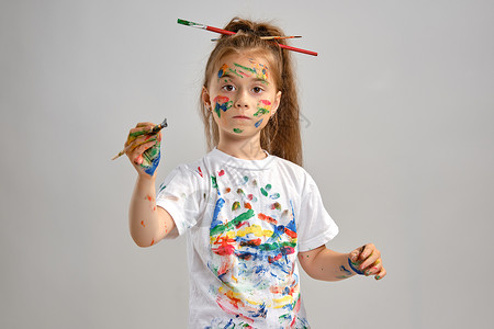身穿白色 T 恤 头发上梳着梳子的小女孩正孤立地站在白色上 用彩绘的手和脸指指点点 特写画架爱好创造力马尾辫工艺艺术品女孩比划女背景图片