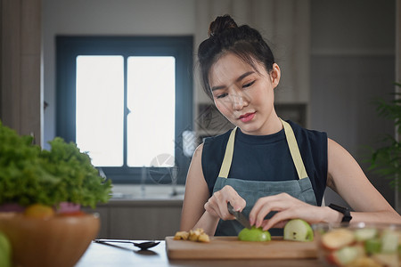 穿着围裙的亚洲女人坐在厨房桌前 在准备健康沙拉时在木制剪板上切苹果图片