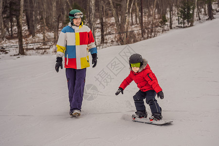 单板滑雪教练教男孩滑雪 冬季儿童活动 儿童冬季运动 生活方式女士寄宿生骑术行动头盔父亲木板活力家庭父母背景