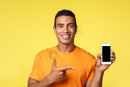 宣传展板展穿着橙色T恤衫的英俊漂亮的年轻男子 手持智能手机移动显示 微笑满意 建议获得应用程序 宣传电话申请 在网上炫耀什么买到的东西等展背景