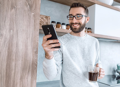 男人灵光一现用智能手机和咖啡杯 站在厨房里的人笑着男人上网杯子互联网细胞眼镜技术饮料电话享受胡须背景