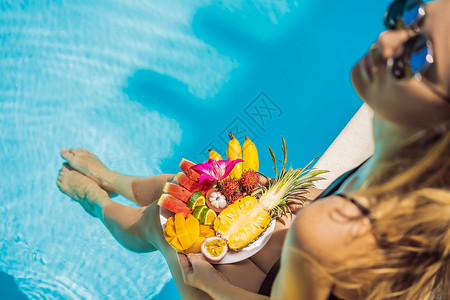 女青年在旅馆泳池旁放松和吃水果盘 夏季食用异样食品 游泳池边的双腿与健康食物相照 上面最美景情调排毒异国比基尼女士水果水池西瓜热背景图片