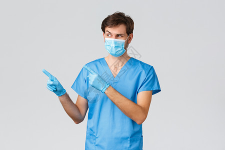 Covid-19 医护人员和医院概念 身着蓝色磨砂膏 手套和医用面具的可疑而多疑的男医生 看起来很好奇 思考着奇怪的宣传片 灰色背景