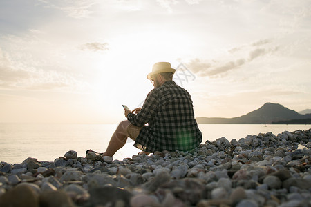 手机视图素材在阳光明媚的日子里 坐在海滩露台上的智能手机男子的后侧视图 背景外有复制空间  假期或自由职业者或社交网络概念男性帽子热带石头日背景