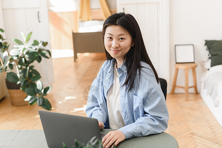 年轻亚洲妇女在家里用笔记本电脑工作 看照相机 自由商业概念 网络横幅背景图片