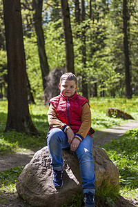 神奇大圆石一个穿着无袖红色夹克的年轻人在春天坐在松树林中的一块巨大的石头上 太阳光线发出的神奇光芒落在男孩身后公园绿色树干植物树桩快乐旅行背景