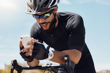 快乐的年轻骑自行车的人戴着头盔和眼镜 同时使用他的智能手机和坐在他的自行车上 适合运动员在户外骑自行车时使用应用程序或在手机上发背景图片